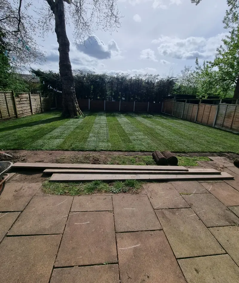 Freshly laid turf in neat stripes across a large back garden, with rolls of turf ready beside the old flags